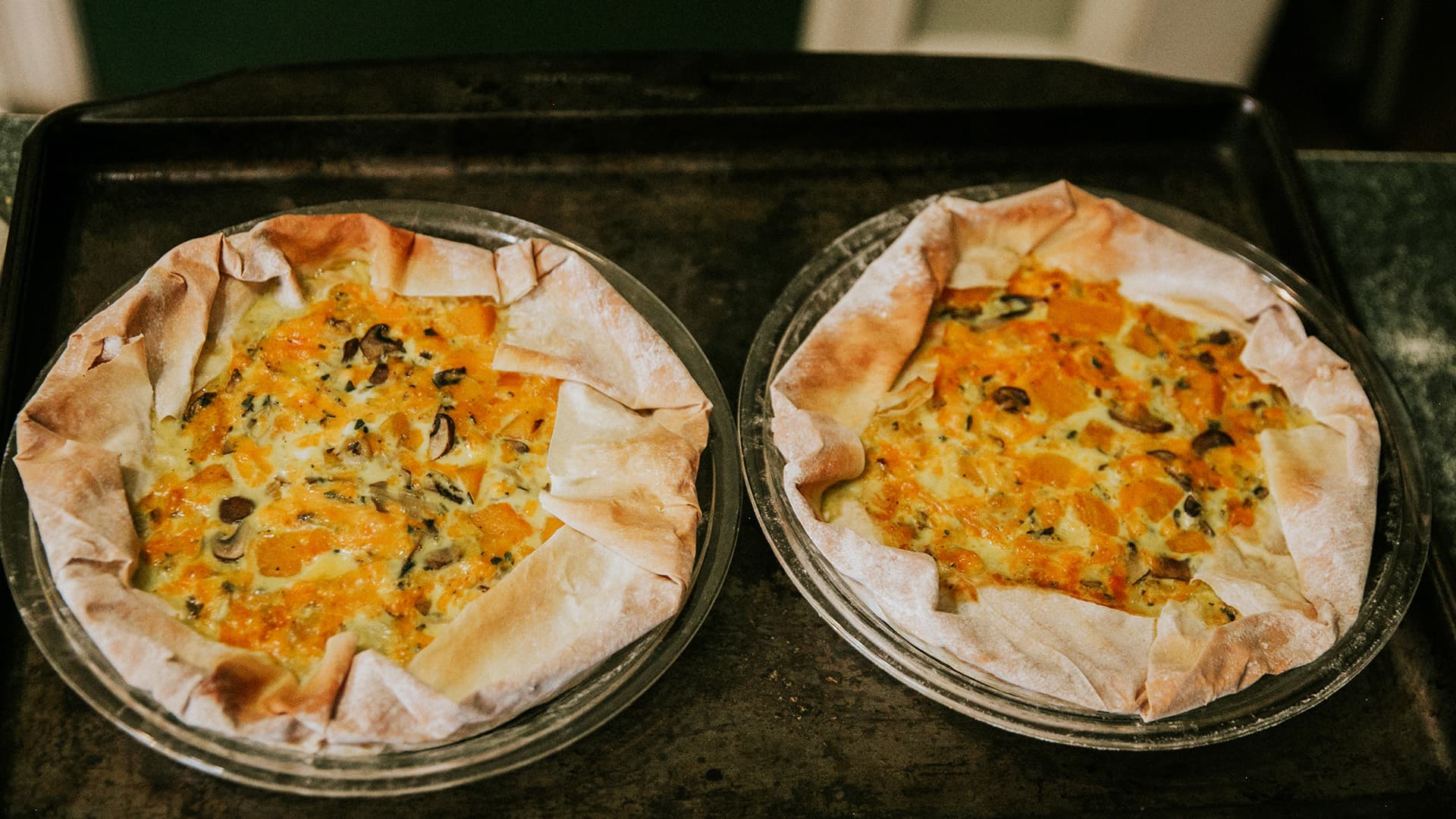 Two baked quiches with golden crusts and colorful fillings on a baking tray.