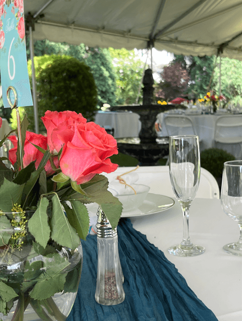 A table set for an outdoor event featuring pink roses, glassware, and a fountain in the background.