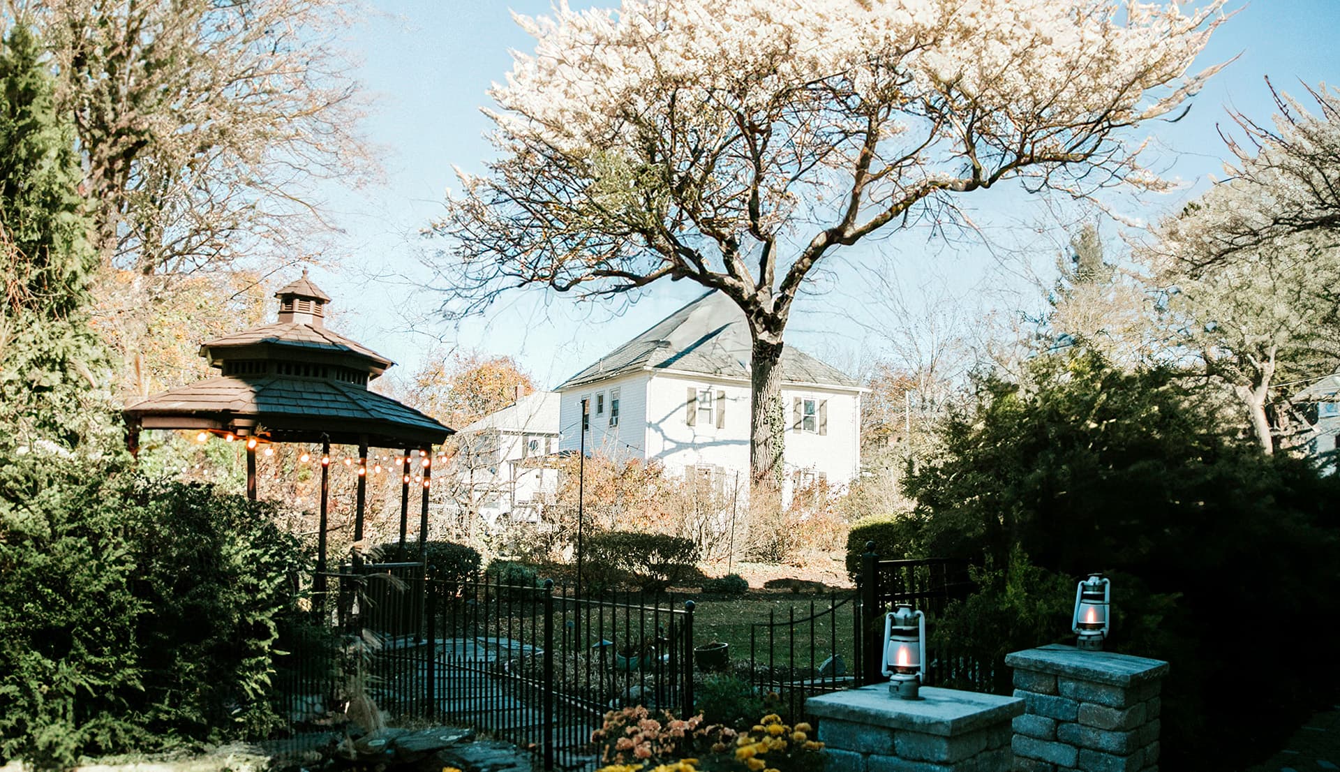 A serene garden scene featuring a gazebo, blossoming tree, and a white house in the background.