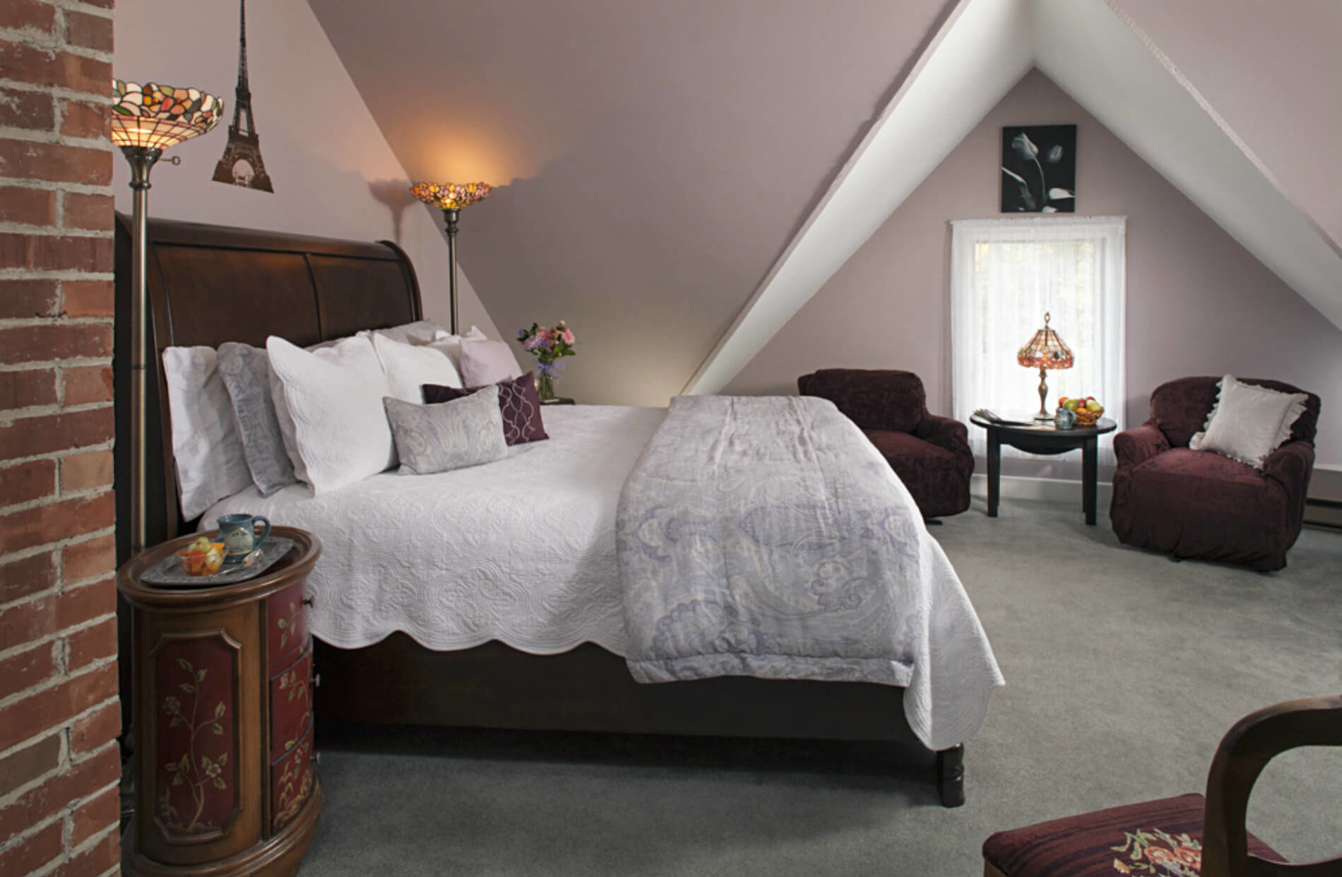 A spacious attic bedroom features a king bed with white and purple bedding, two armchairs, and sloped ceilings with decorative lighting.