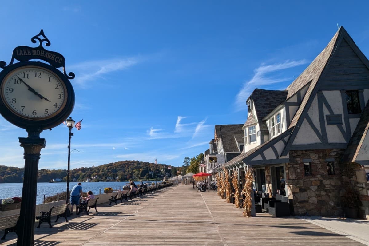 A boardwalk along a lake on a clear day, featuring shops, benches, and a vintage clock.