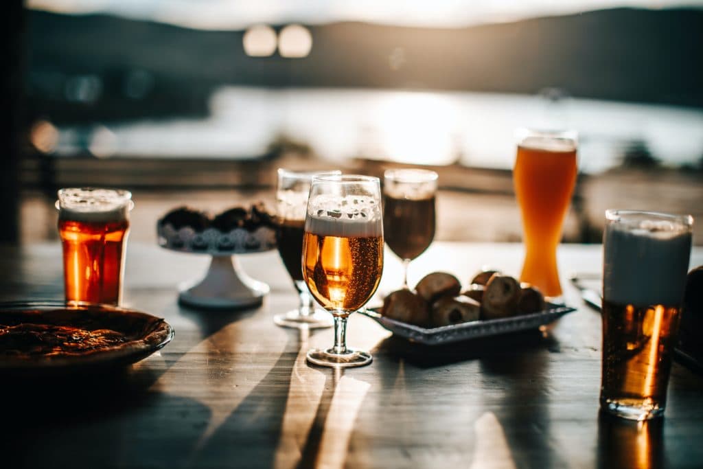 Several glasses of beer and trays of food on a table overlooking a body of water.