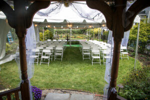 A photograph of the garden set up for a micro wedding. Rows of lawn chairs are carefully placed under a white canopy with soft lighting overhead. In the foreground of the image, the edges of an arch can be seen where the bride & groom will stand