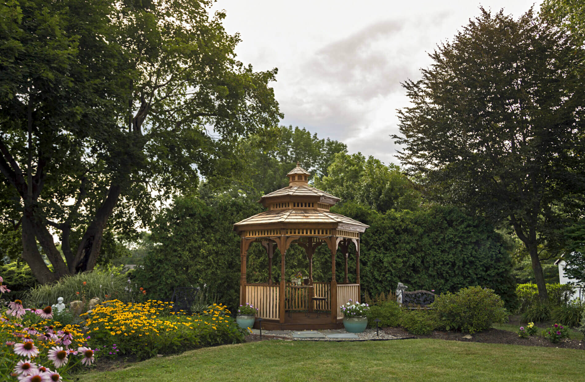 Wooden gazebo surrounded by green trees and lawn with purple and yellow flowers