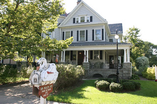 Large Victoria home with wrap-around porch, surrounded by trees.