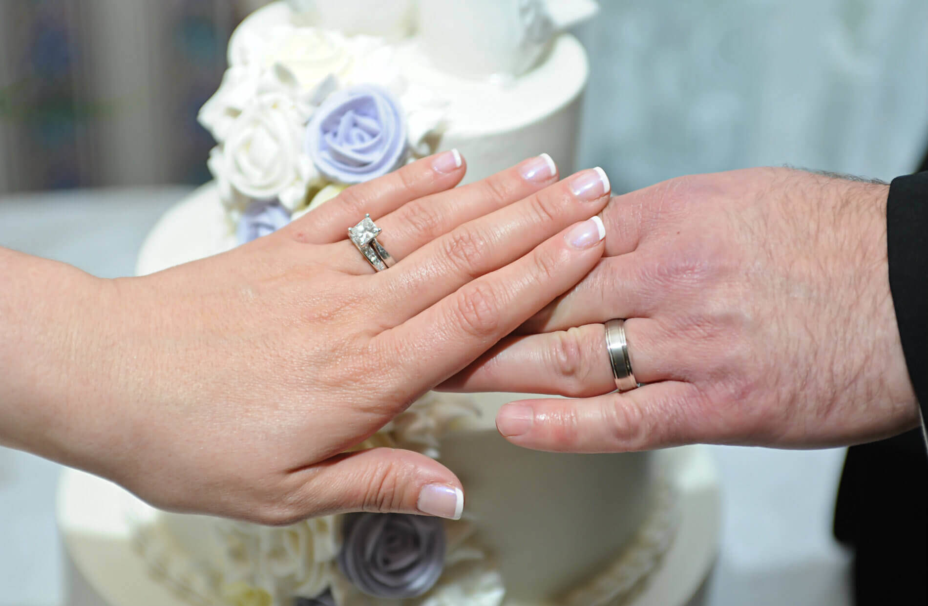 Hands of bride and groom with silver wedding rings in front of white two tier cake with lavender roses