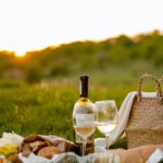 A sunset picnic setup with wine and two glasses over a blanket in the grass.