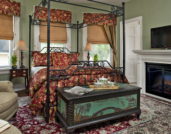 Large 4 post bed in red and yellow bedding on oriental rug in room with two windows