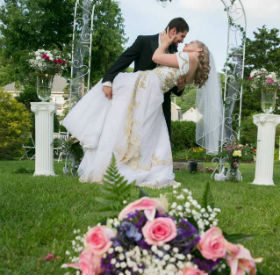 Bride and groom in an embrace. Foreground is a pink rose and baby's breath bouquet. A green arch and two plinths are in the background