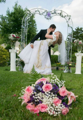 Bride and groom in an embrace. Foreground is a pink rose and baby's breath bouquet. A green arch and two plinths are in the background