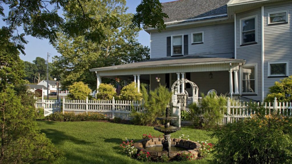 The front of the Whistling Swan Inn, with a white picket fence, lush greenery, and a wraparound porch.